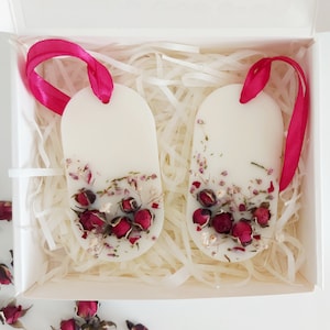 Peut inclure: Deux fondants de cire blancs en forme ovale avec des fleurs séchées et des accents de ruban rose. Les fondants sont dans une boîte blanche avec du papier blanc déchiqueté.