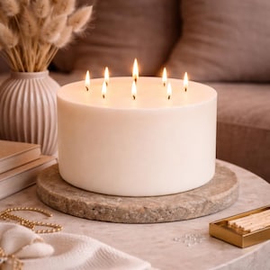Peut inclure: Une grande bougie ivoire avec sept mèches allumées. La bougie repose sur une base ronde en forme de pierre. Un vase avec des fleurs séchées et une boîte d'allumettes dorée sont en arrière-plan, suggérant un thème de décoration intérieure.