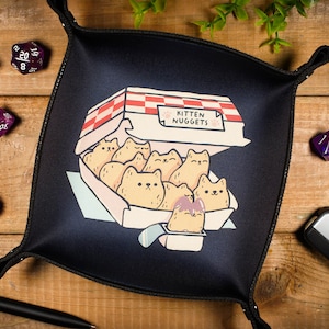 Könnte beinhalten: Ein schwarzes Würfeltablett mit weißem Rand. Das Tablett zeigt eine Comic-Illustration einer Schachtel mit Katzen-Nuggets. Die Schachtel ist geöffnet und enthält mehrere Katzen-Nuggets. Der Text "KITTEN NUGGETS" ist auf der Schachtel aufgedruckt. Neben dem Tablett liegen mehrere lila Würfel auf einer Holzoberfläche.