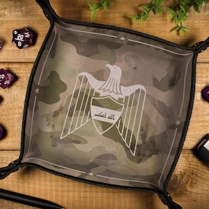 May include: A camouflage-patterned dice tray with a white eagle emblem and Arabic text, surrounded by several purple dice. The tray is on a wooden surface, with a pen and car key fob visible.