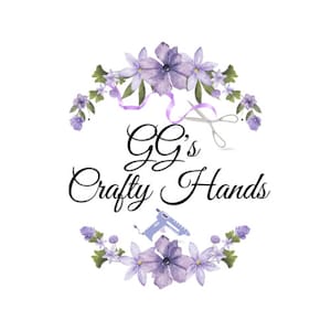 K&ouml;nnte beinhalten: Ein Logo f&uuml;r ein Handwerksgesch&auml;ft namens "GG's Crafty Hands". Das Logo zeigt einen Kranz aus lila Blumen, eine Schere und eine Klebepistole.