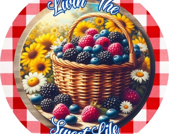 May include: Round image featuring a woven basket overflowing with fresh berries: raspberries, blueberries, and blackberries. The basket sits on a wooden surface, surrounded by sunflowers and daisies. The text "Livin' The Sweet Life" is arched above and below the basket, set against a red and white checkered background.