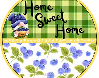 May include: A round sign featuring a green plaid background with the text Home Sweet Home in black. A gnome wearing a blue hat holds a basket of blueberries. The bottom section displays a white background with a pattern of blueberries and green leaves.