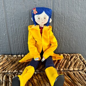 May include: A handmade doll with blue yarn hair, wearing a yellow raincoat, blue jeans, and yellow rain boots. The doll is sitting on a woven wicker basket.