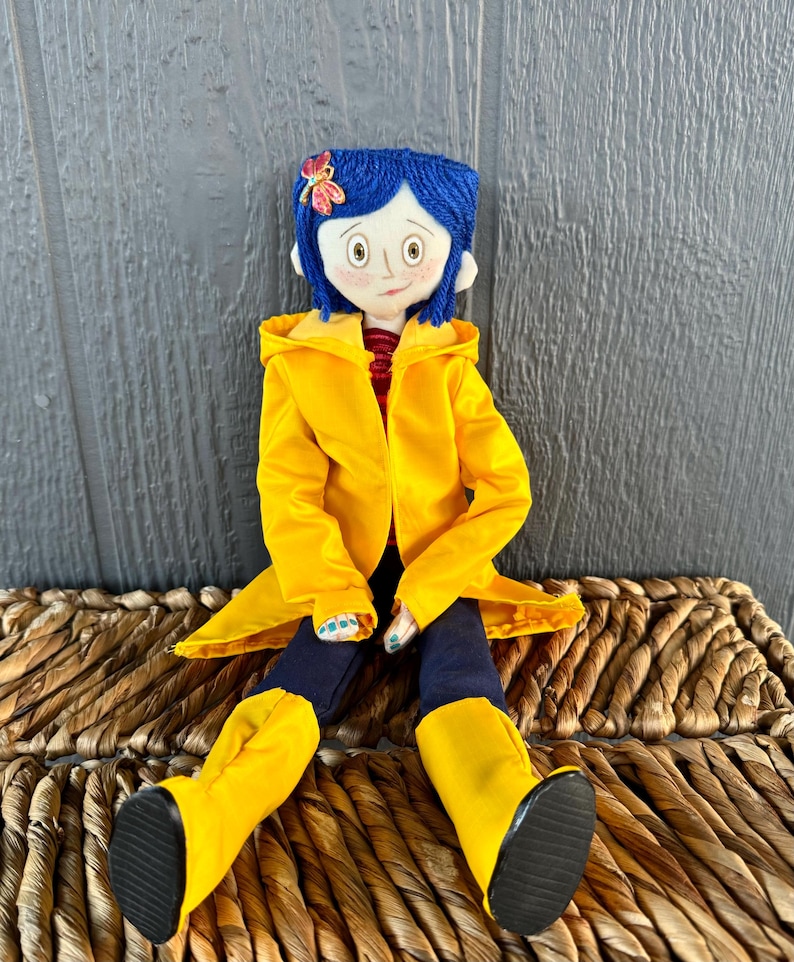 May include: A handmade doll with blue yarn hair, wearing a yellow raincoat, blue jeans, and yellow rain boots. The doll is sitting on a woven wicker basket.