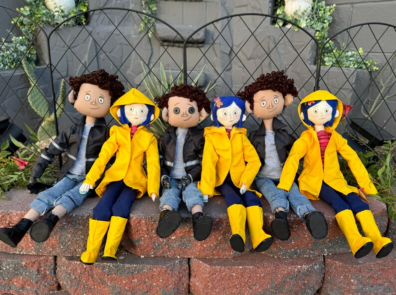 May include: Five plush dolls, including three wearing yellow raincoats, sitting on a brick wall. The dolls have black hair, brown eyes, and are wearing jeans and jackets.