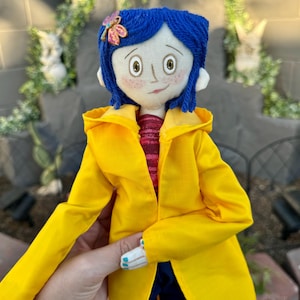 May include: A handmade doll with blue yarn hair, a yellow raincoat, and a red and white striped shirt. The doll is wearing a flower-shaped brooch with pink and purple accents.