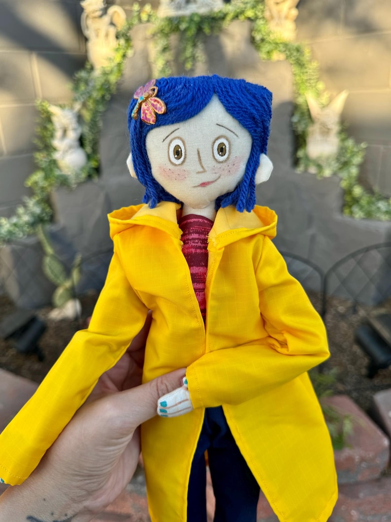 May include: A handmade doll with blue yarn hair, a yellow raincoat, and a red and white striped shirt. The doll is wearing a flower-shaped brooch with pink and purple accents.