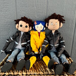 May include: Three handmade fabric dolls, two with brown hair and black leather jackets, and one with blue hair wearing a yellow raincoat. The dolls are sitting on a woven basket.