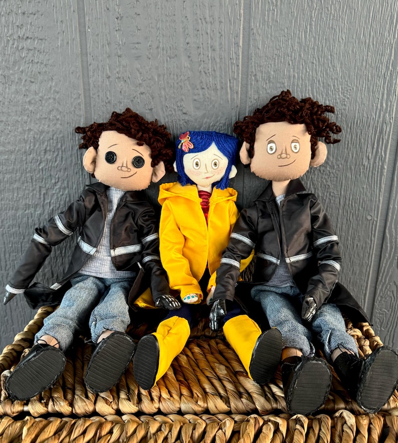 May include: Three handmade fabric dolls, two with brown hair and black leather jackets, and one with blue hair wearing a yellow raincoat. The dolls are sitting on a woven basket.