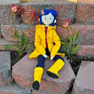 May include: A handmade doll wearing a yellow raincoat, blue jeans, and yellow rain boots. The doll has blue hair, a blue flower in her hair, and a painted face.