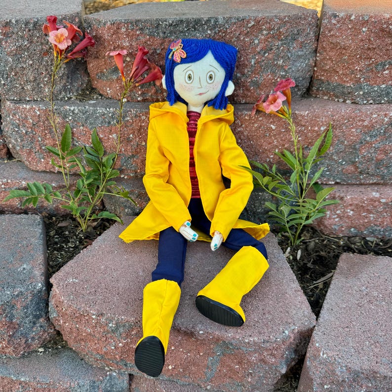 May include: A handmade doll wearing a yellow raincoat, blue jeans, and yellow rain boots. The doll has blue hair, a blue flower in her hair, and a painted face.