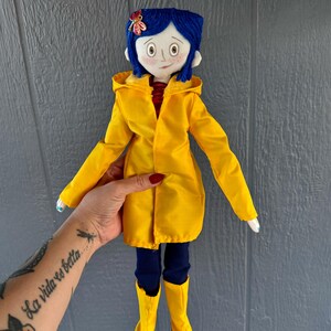 May include: A handmade doll wearing a yellow raincoat, blue jeans, and yellow rain boots. The doll has blue yarn hair and a red flower on her head.