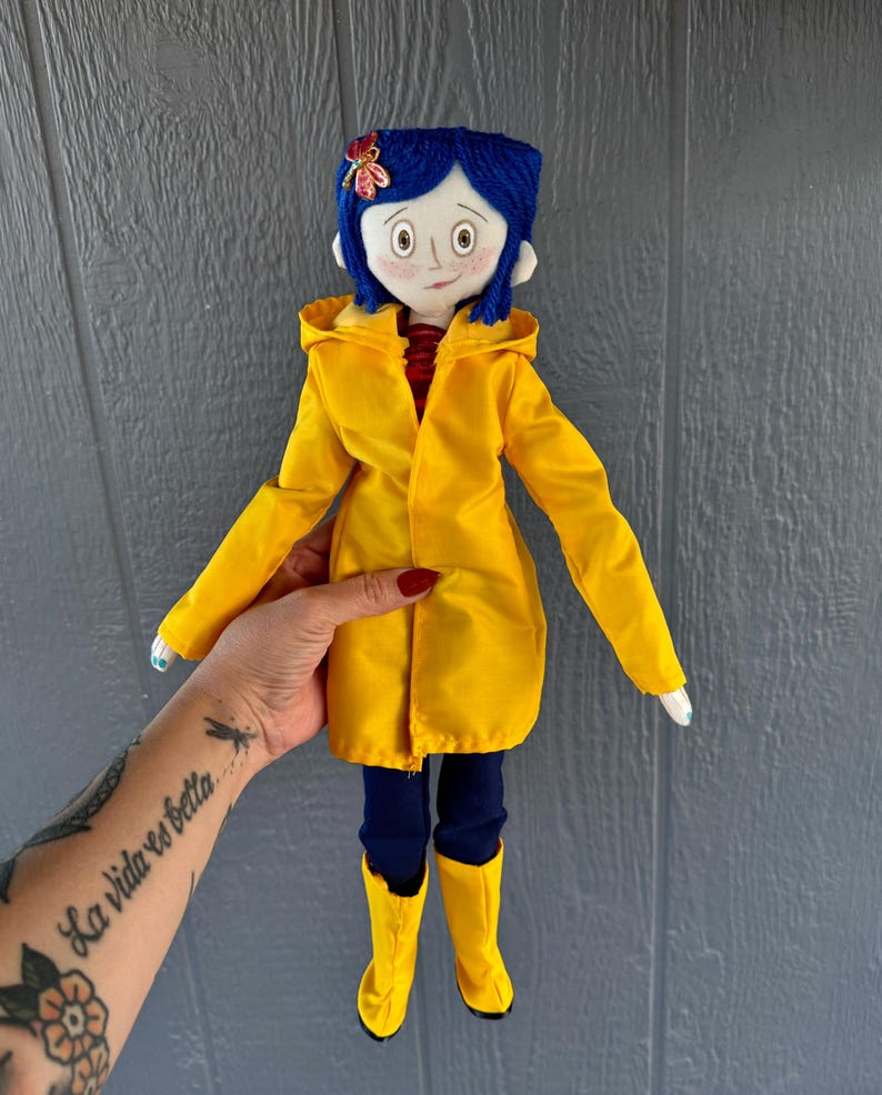 May include: A handmade doll wearing a yellow raincoat, blue jeans, and yellow rain boots. The doll has blue yarn hair and a red flower on her head.