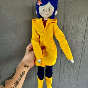 May include: A handmade doll with blue hair, wearing a yellow raincoat, black pants, and yellow boots. The doll has a painted face with large eyes and a red mouth.
