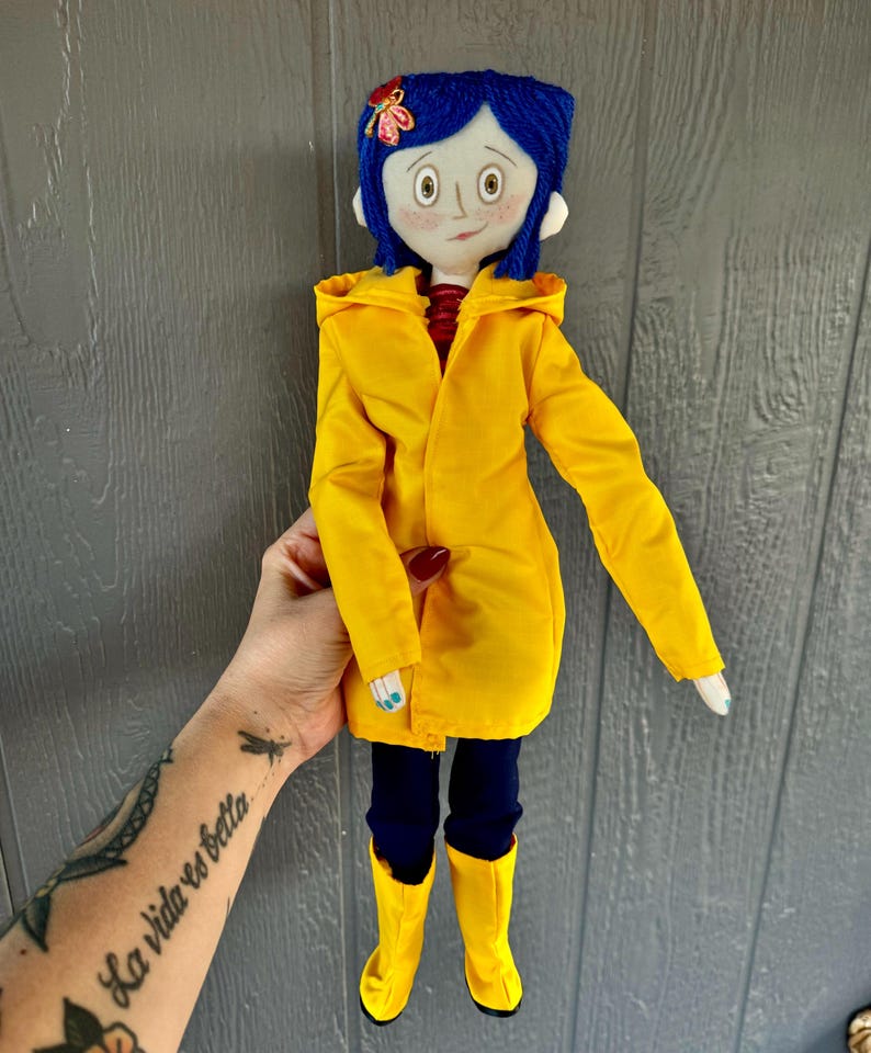 May include: A handmade doll with blue hair, wearing a yellow raincoat, black pants, and yellow boots. The doll has a painted face with large eyes and a red mouth.