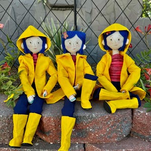 May include: Three handmade dolls dressed in yellow raincoats and blue jeans. The dolls have blue hair, brown eyes, and are wearing yellow rain boots.