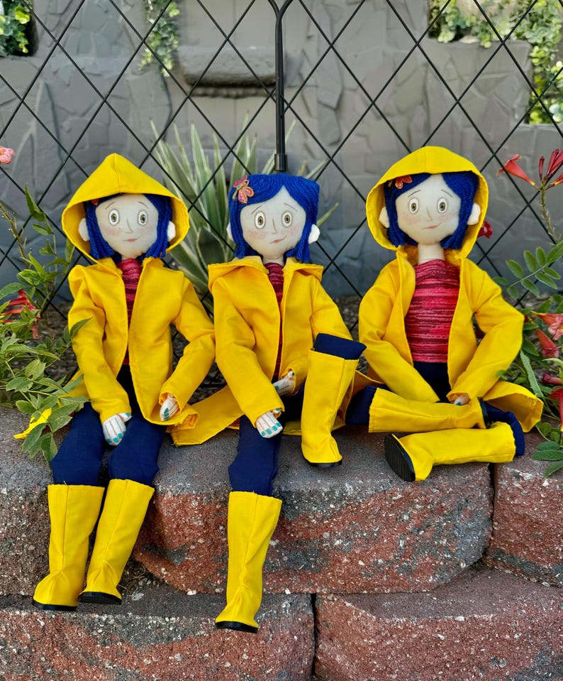 May include: Three handmade dolls dressed in yellow raincoats and blue jeans. The dolls have blue hair, brown eyes, and are wearing yellow rain boots.