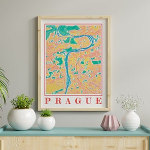 Op de afbeelding: Een ingelijste print van een kleurrijke kaart van Praag, Tsjechië. De kaart is in een geel, groen en rood kleurenschema. De rivier van de stad is in blauw weergegeven. Het woord "PRAG" staat in rood onderaan de kaart.