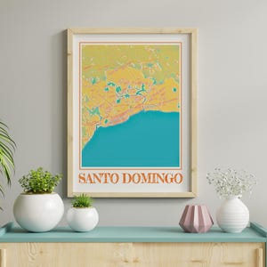 May include: A framed print of a map of Santo Domingo, Dominican Republic. The map is in a vintage style with a yellow, orange, and green colour scheme. The print is framed in a light wood frame and is hanging on a white wall.