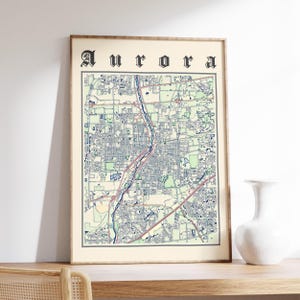 May include: Framed map print of Aurora, showing a detailed city layout with streets, parks, and waterways. The map is in shades of blue, green, and beige, with the city name in a gothic font at the top. The print is in a wooden frame.