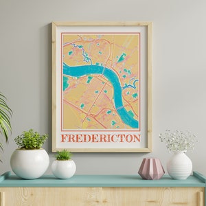 May include: A framed print of a map of Fredericton, New Brunswick, Canada. The map is in a retro style with a yellow background and blue river. The city name is printed in red at the bottom of the map.