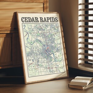 May include: A framed map of Cedar Rapids, Iowa, with a vintage style design. The map is printed on cream-colored paper with blue and red lines and text. The frame is brown wood.