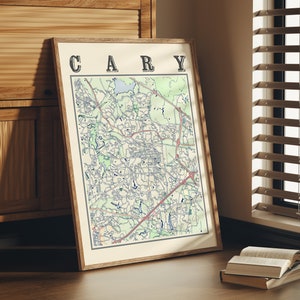 May include: A framed map of Cary, North Carolina, with the city name printed in bold letters at the top. The map is in a light wood frame and is hanging on a wall.