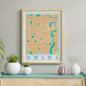 Puede incluir: Una impresión enmarcada de un mapa de Omaha, Nebraska. El mapa es de estilo vintage con un fondo amarillo y naranja y líneas verdes. La palabra "OMAHA" está impresa en azul en la parte inferior del mapa.