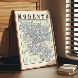 May include: A framed map of Modesto, California, with the city name printed in bold black letters at the top. The map is in a light wood frame and is hanging on a wall.