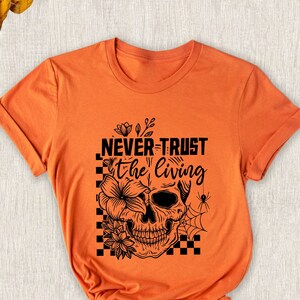 May include: Orange t-shirt with the text "NEVER TRUST the LIVING" above a skull graphic. The skull is decorated with flowers and a spider web. The shirt is folded and has rolled-up sleeves.