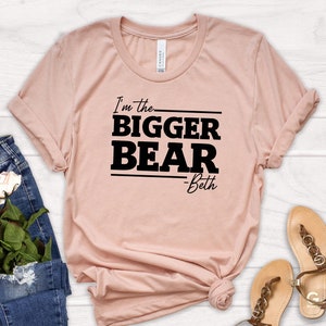 May include: A light pink t-shirt with black text that reads "I'm the BIGGER BEAR -Beth". The shirt is laid out on a white wooden surface with a pair of blue jeans and sandals.