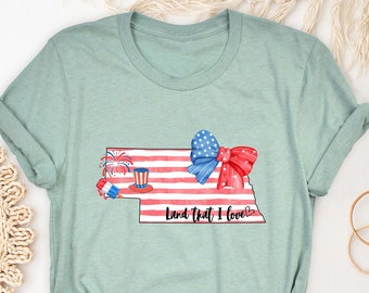 May include: A light blue t-shirt with a watercolor design of the state of Nebraska. The design features a red, white, and blue American flag, a red bow, fireworks, and a red, white, and blue ice cream cone. The text "Land That I Love" is written below the design.