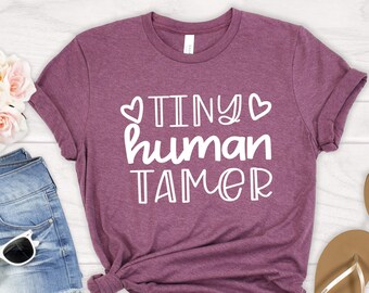 May include: A maroon t-shirt with the text "TINY human TAMER" in white with two small hearts on either side of the word "TINY".
