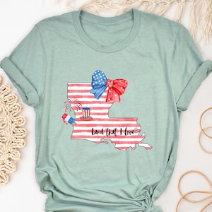 K&ouml;nnte beinhalten: Ein hellblaues T-Shirt mit einem Aquarell-Design des Bundesstaates Louisiana. Das Design zeigt eine rot-wei&szlig;-blaue Schleife, Feuerwerk und den Text "Land That I Love".