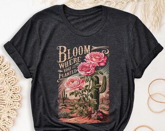 May include: Dark grey heather t-shirt with a vintage-style graphic of pink flowers and a cactus. The graphic says "Bloom Where You Are Planted".
