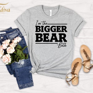 May include: Gray t-shirt with black text that reads "I'm the bigger bear - Beth". The shirt is laid flat on a wooden surface with a pair of blue jeans and a bouquet of pink roses.