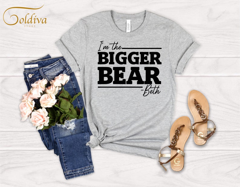May include: Gray t-shirt with black text that reads "I'm the bigger bear - Beth". The shirt is laid flat on a wooden surface with a pair of blue jeans and a bouquet of pink roses.