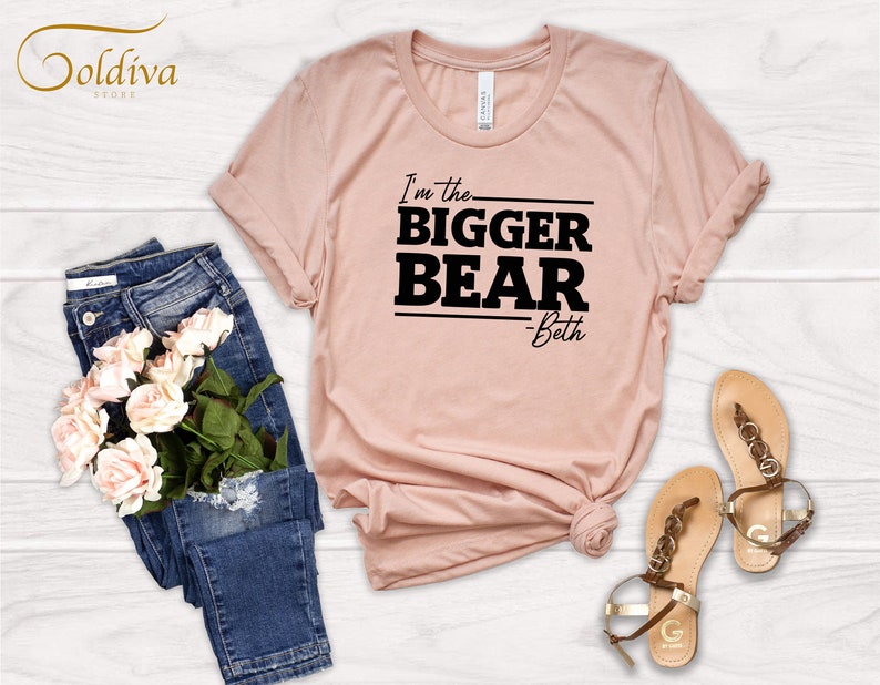 May include: A light pink t-shirt with black text that reads "I'm the BIGGER BEAR -Beth". The shirt is laid out on a white wooden surface with a pair of blue jeans and sandals.