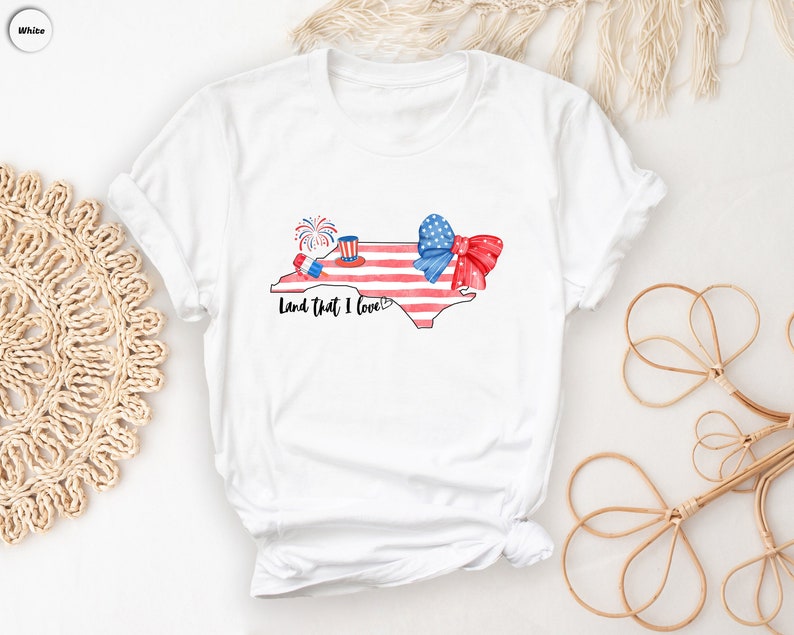 K&ouml;nnte beinhalten: Wei&szlig;es T-Shirt mit einer Aquarell-Illustration des Bundesstaates North Carolina. Der Bundesstaat ist mit roten, wei&szlig;en und blauen Sternen und Streifen, einer roten Schleife und Feuerwerk verziert. Der Text "Land that I love" ist unter dem Bundesstaat gedruckt.