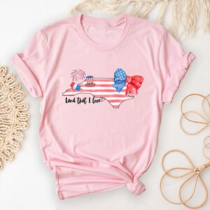 K&ouml;nnte beinhalten: Rosa T-Shirt mit einem Aquarell-Design des Bundesstaates North Carolina, einer rot-wei&szlig;-blauen Schleife und Feuerwerk. Der Text "Land that I love" ist unter dem Design gedruckt.