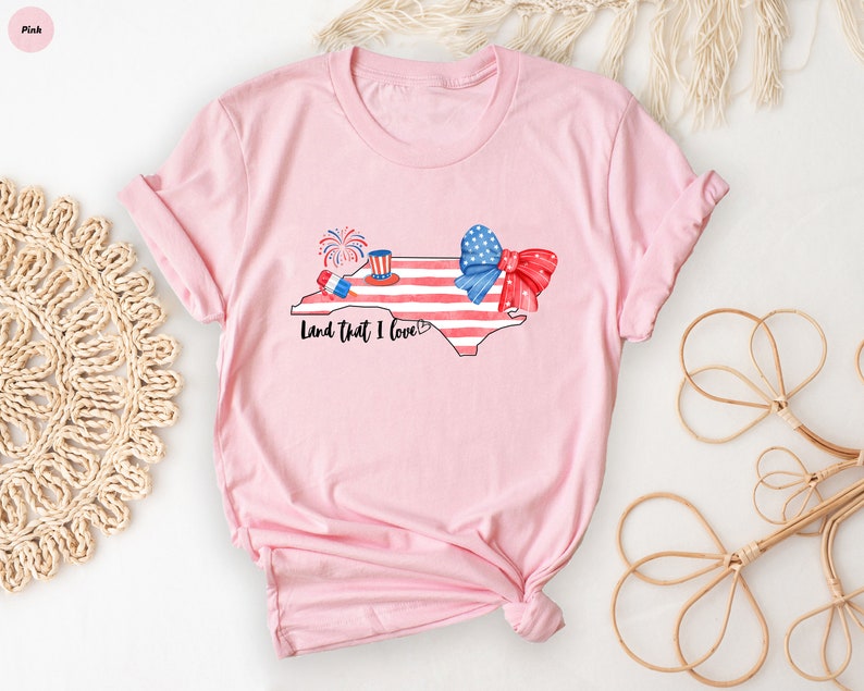 K&ouml;nnte beinhalten: Rosa T-Shirt mit einem Aquarell-Design des Bundesstaates North Carolina, einer rot-wei&szlig;-blauen Schleife und Feuerwerk. Der Text "Land that I love" ist unter dem Design gedruckt.