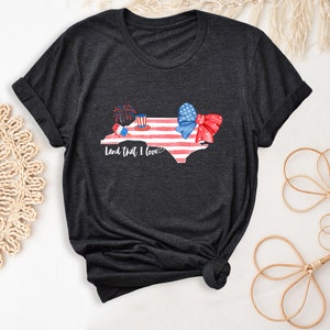K&ouml;nnte beinhalten: Dunkelgraues T-Shirt mit einem Aquarell-Motiv des Bundesstaates North Carolina. Der Bundesstaat ist mit roten, wei&szlig;en und blauen Sternen und Streifen, einer roten Schleife und einem roten, wei&szlig;en und blauen Zylinder mit Feuerwerk verziert. Der Text "Land that I love!" ist unter dem Bundesstaat gedruckt.