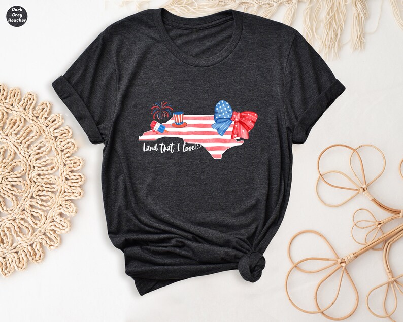 K&ouml;nnte beinhalten: Dunkelgraues T-Shirt mit einem Aquarell-Motiv des Bundesstaates North Carolina. Der Bundesstaat ist mit roten, wei&szlig;en und blauen Sternen und Streifen, einer roten Schleife und einem roten, wei&szlig;en und blauen Zylinder mit Feuerwerk verziert. Der Text "Land that I love!" ist unter dem Bundesstaat gedruckt.