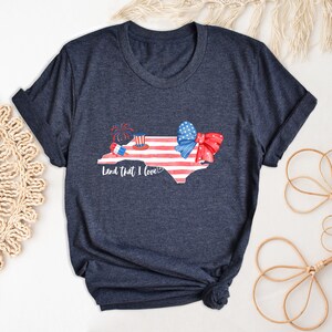 K&ouml;nnte beinhalten: Ein marineblaues T-Shirt mit einem Aquarell-Design des Bundesstaates North Carolina. Der Bundesstaat ist mit roten, wei&szlig;en und blauen Sternen und Streifen, einer roten Schleife und Feuerwerk verziert. Der Text "Land that I love" ist unter dem Bundesstaat gedruckt.