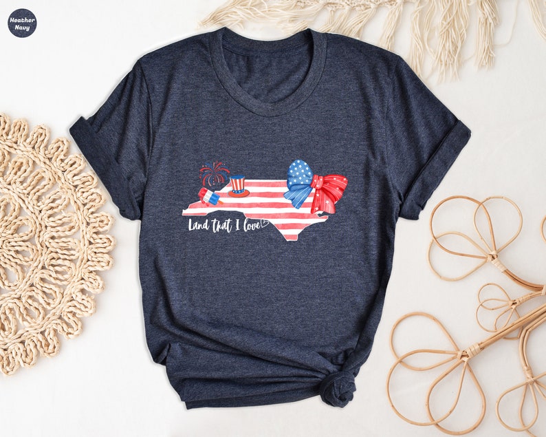 K&ouml;nnte beinhalten: Ein marineblaues T-Shirt mit einem Aquarell-Design des Bundesstaates North Carolina. Der Bundesstaat ist mit roten, wei&szlig;en und blauen Sternen und Streifen, einer roten Schleife und Feuerwerk verziert. Der Text "Land that I love" ist unter dem Bundesstaat gedruckt.