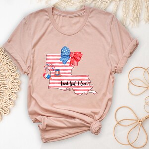 K&ouml;nnte beinhalten: Ein T-Shirt in der Farbe Heidepfirsich mit einem Aufdruck des Bundesstaates Louisiana. Der Aufdruck zeigt eine rot-wei&szlig;-blaue amerikanische Flagge, eine rote Schleife und Feuerwerk. Der Text "Land That I Love" steht unter dem Aufdruck.