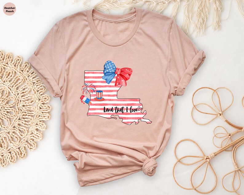 K&ouml;nnte beinhalten: Ein T-Shirt in der Farbe Heidepfirsich mit einem Aufdruck des Bundesstaates Louisiana. Der Aufdruck zeigt eine rot-wei&szlig;-blaue amerikanische Flagge, eine rote Schleife und Feuerwerk. Der Text "Land That I Love" steht unter dem Aufdruck.