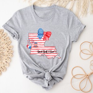 K&ouml;nnte beinhalten: Graues T-Shirt mit einem Aufdruck des Bundesstaates Louisiana. Der Bundesstaat ist mit roten, wei&szlig;en und blauen Streifen, einer roten, wei&szlig;en und blauen Schleife und Feuerwerk verziert. Der Text "Land that I love" steht unter dem Bundesstaat.