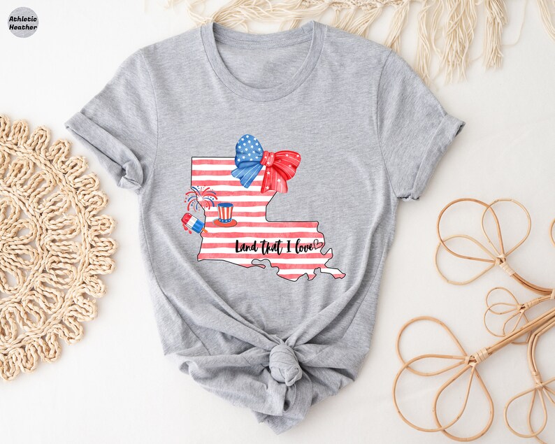 K&ouml;nnte beinhalten: Graues T-Shirt mit einem Aufdruck des Bundesstaates Louisiana. Der Bundesstaat ist mit roten, wei&szlig;en und blauen Streifen, einer roten, wei&szlig;en und blauen Schleife und Feuerwerk verziert. Der Text "Land that I love" steht unter dem Bundesstaat.